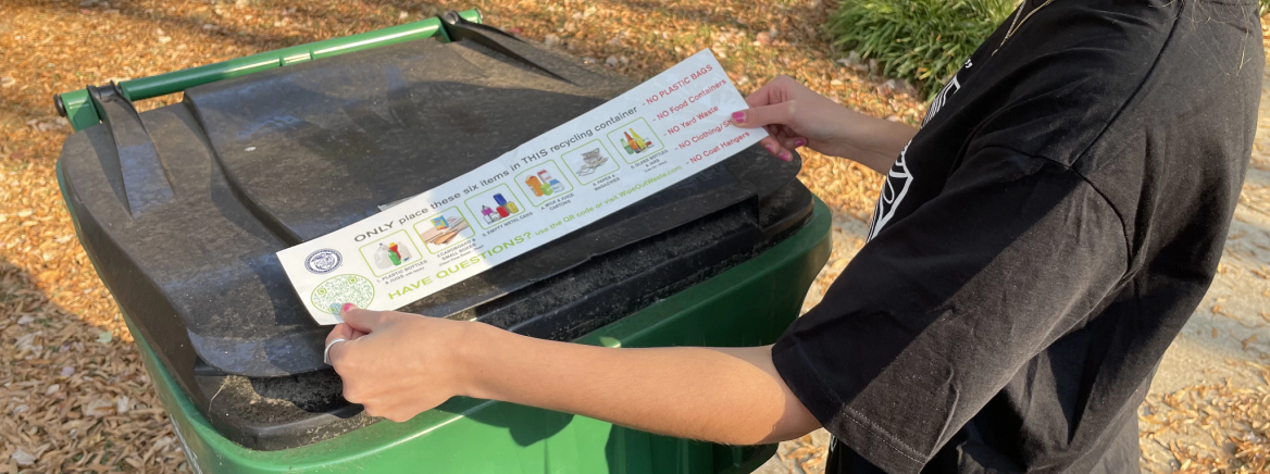 A person adheres to a recycling cart a sticker that lists recyclable items.