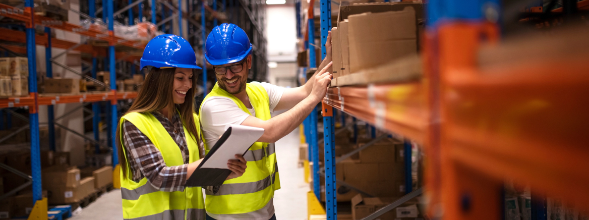 Two employees in a warehouse A female and male employee wearing yellow construction vests and blue hard hats landing in a warehouse and looking at a notepad.