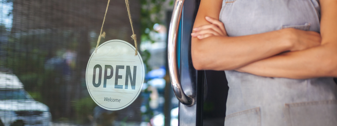 Shop worker next to a storefront&#039;s &quot;Open&quot; sign.