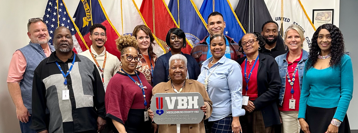 Veterans Bridge Home Janene Stewart McGee Provider of the Year Award Mecklenburg County Veterans Services employees stand in front of flags holding the Veterans Bridge Home Janene Stewart McGee Provider of the Year award.