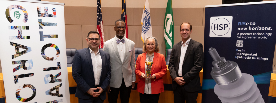 HSP, BOCC Chair and Mayor HSP executives stand with Chair Mark Jerrell and Charlotte Mayor Vi Lyles in the meeting chamber of the Government Center. On their left is a banner that reads Charlotte, on the right a banner reading HSP.