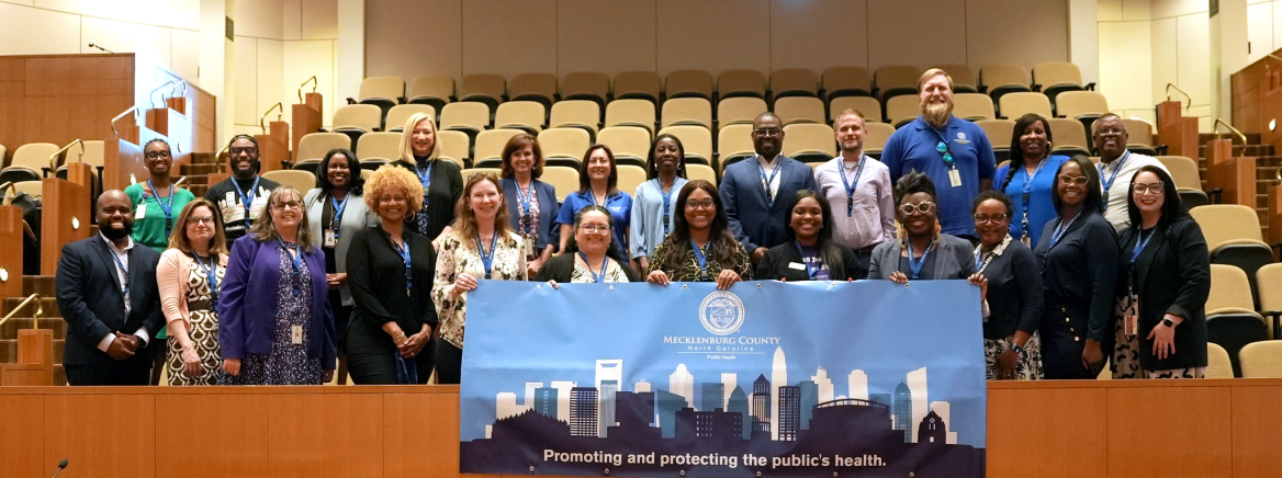 Public Health Group Photo in CMGC Chamber Dr. Raynard Washington and members of Public Health stand in the first two rows of the CMGC meeting chamber holding a sign that says promoting and protecting the public's health.