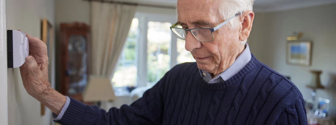 Worried Senior Man With Bill Turning Down Central Heating Thermostat At Home In Energy Crisis