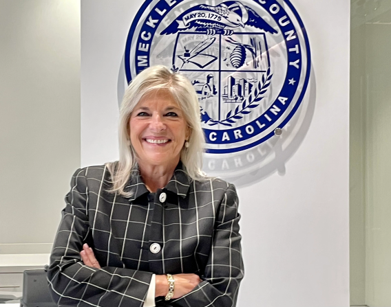 Dena R. Diorio wearing a Black Checkered blazer standing in front of the County seal.